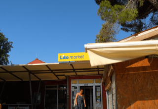 Vrouw verlaat Lito Market op Camping Polari in Istrië, Kroatië, met blauwe lucht en bomen achter haar.