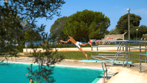Niño salta desde el trampolín a la piscina en Camping Polari, Istria, Croacia, rodeado de árboles y naturaleza.