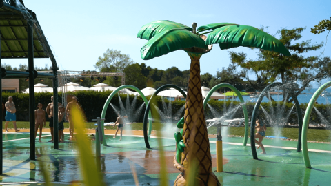 Niños jugando en un parque acuático con fuente de palmera en Camping Polari, parque de vacaciones en Istria, Croacia.
