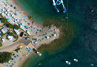 Vista aerea di Camping Krk in Croazia con spiaggia, camper, ombrelloni e barche sull’acqua limpida.