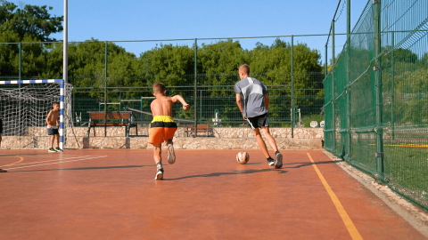 Kids play football on an outdoor court surrounded by greenery at Camping Krk in Primorje-Gorski Kotar, Croatia.