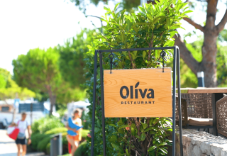 Insegna di legno del ristorante Oliva presso Camping Krk, circondata da piante verdi, in Croazia.