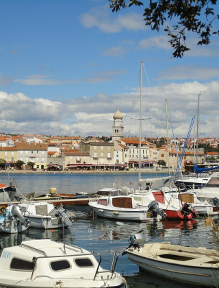 Vista de barcos atracados en un puerto pintoresco, con edificios de tejados rojos y torre de iglesia al fondo.