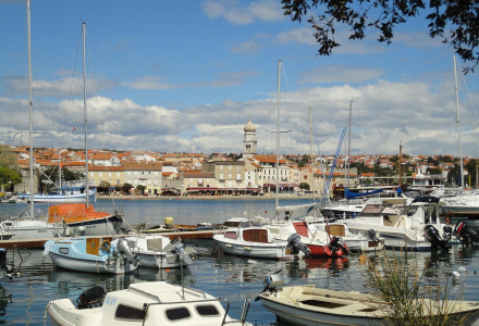 Vista de barcos atracados en un puerto pintoresco, con edificios de tejados rojos y torre de iglesia al fondo.