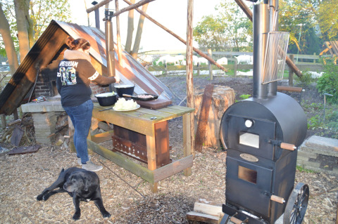 Una mujer cocina al aire libre en un área rústica con horno negro y un perro, rodeados de naturaleza y árboles.