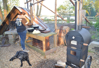 Una mujer cocina al aire libre en un área rústica con horno negro y un perro, rodeados de naturaleza y árboles.