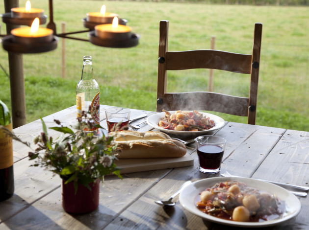 Buitenmaaltijd op houten tafel met wijn, brood, warme stoofpot en kaarsen, met uitzicht op Duitse velden.