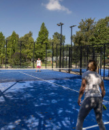 Visitantes del parque vacacional juegan pádel en una pista azul al aire libre rodeada de malla y árboles en Camping Papillon.