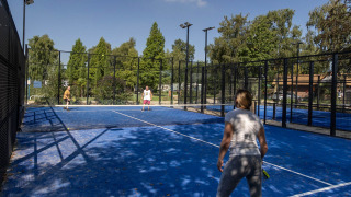 Visitantes del parque vacacional juegan pádel en una pista azul al aire libre rodeada de malla y árboles en Camping Papillon.