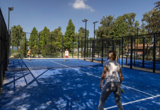 Vakantiegangers spelen padel op het blauwe buitenveld met omheining en bomen bij Camping Papillon in Limburg.