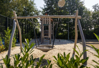 Legeplads med gynger og rutsjebane på Camping Papillon i Belgisk Limburg, omgivet af træer og sand.