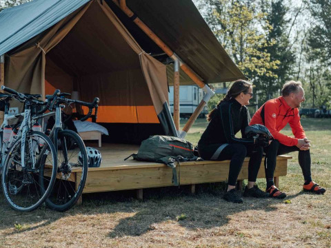 Dos ciclistas con ropa deportiva se sientan frente a una tienda Safari tent XS con bicicletas y mochilas.