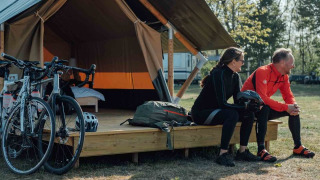 Dos ciclistas con ropa deportiva se sientan frente a una tienda Safari tent XS con bicicletas y mochilas.