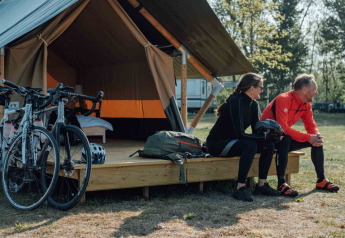 Dos ciclistas con ropa deportiva se sientan frente a una tienda Safari tent XS con bicicletas y mochilas.