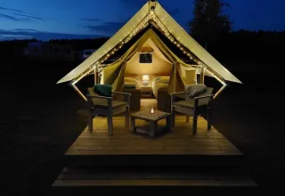 Tienda safari XS en Camp Kyllburg, Alemania, iluminada por la noche con luces y asientos acogedores afuera.