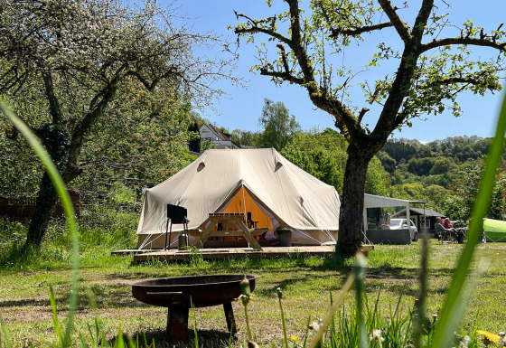 Glamping-Platz mit Tipi-Zelt, umgeben von grünen Bäumen und Wiese bei strahlend blauem Himmel.
