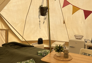 Binnenkant van een Tipi-tent met bed, eettafel, koelkast, kussens en kleurrijke vlaggetjes als decor.