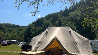 Grote tipi tent op houten terras met picknickbank, omgeven door groene heuvels en een blauwe lucht.