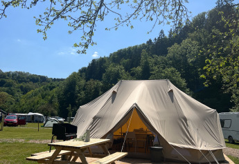 Stort tipitelt på træterrasse med picnicbord, omgivet af grønne bakker og campingvogne under blå himmel.