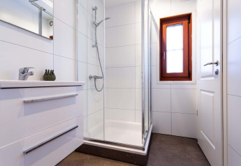 Baño moderno con ducha de vidrio, azulejos blancos, lavabo, espejo y ventana en una cabaña.