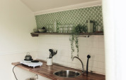 Pequeña cocina en una cabaña con azulejos verdes, encimera de madera y estante abierto con plantas.