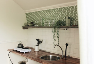 Pequeña cocina en una cabaña con azulejos verdes, encimera de madera y estante abierto con plantas.