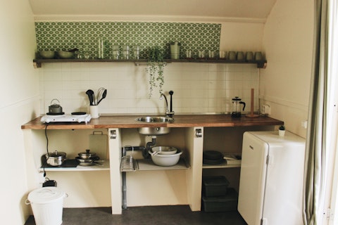 Cocina rústica en una cabaña con estantes abiertos, azulejos verdes, nevera pequeña y utensilios de cocina.