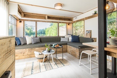 Lyst loungeområde i en lodge med store vinduer, træbænk, pyntepuder og små borde med planter.