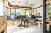 Lyst loungeområde i en lodge med store vinduer, træbænk, pyntepuder og små borde med planter.