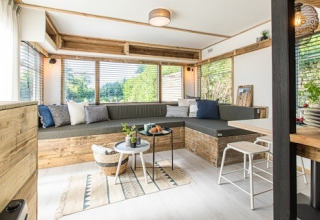 Sala luminosa en una cabaña, con amplios ventanales, sofá de madera, cojines y mesas modernas.