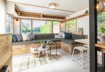 Salotto luminoso in un lodge con grandi finestre, divano in legno, cuscini e tavolini moderni.