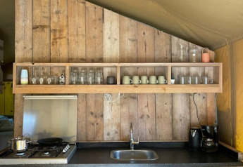 Rustic kitchen in safari tent at 7Huizen aan Zee, Netherlands, with wood panel walls and open shelving.
