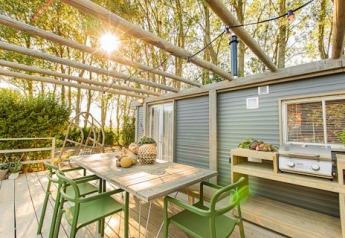 Udendørs terrasse ved Lodge 2 og 3 med spisebord, grønne stole og grill, omgivet af skov og sollys.