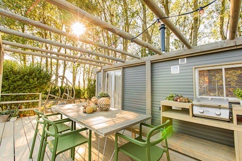 Terrasse von Lodge 2 und 3 mit Holztisch, grünen Stühlen, Grill und Sonnenlicht im Wald.