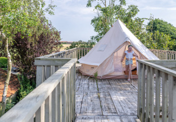 Et barn træder ud af et telt placeret på en trætarrasse omgivet af grønne træer ved Lodge 2 og 3, 7Huizen aan Zee, Holland.