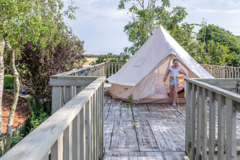 Et barn træder ud af et telt placeret på en trætarrasse omgivet af grønne træer ved Lodge 2 og 3, 7Huizen aan Zee, Holland.
