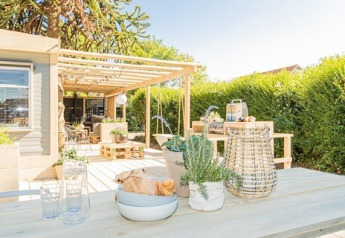 Terraza soleada con mesa de madera, plantas y farol en Lodge 1 y 4 en 7Huizen aan Zee, Países Bajos.