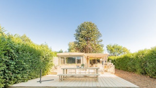 Große Holzterrasse mit Tisch und Bank vor Lodge 1 und 4, umgeben von Hecken und strahlend blauem Himmel.
