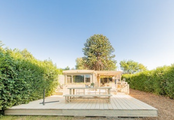 Große Holzterrasse mit Tisch und Bank vor Lodge 1 und 4, umgeben von Hecken und strahlend blauem Himmel.