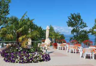 Udendørs terrasse med blomster, palmer, borde og udsigt over søen på Camping Cisano/San Vito i Veneto.