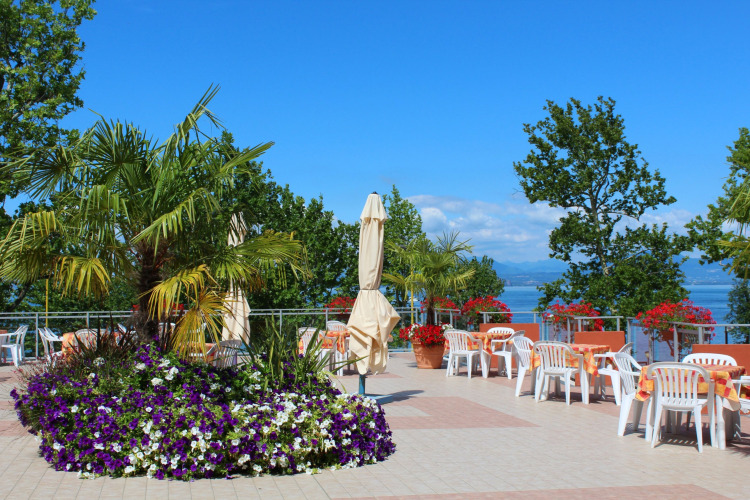 Outdoor terrace at Camping Cisano/San Vito featuring plastic chairs, colorful flowers, and a lake view in Italy.