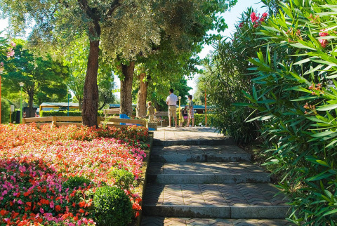 En solrig sti i Camping Cisano/San Vito, Veneto, Italien, omgivet af blomster og træer med mennesker i det fjerne.
