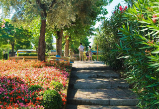 Un sentiero soleggiato al Camping Cisano/San Vito in Veneto, Italia, circondato da fiori e alberi, con persone sullo sfondo.