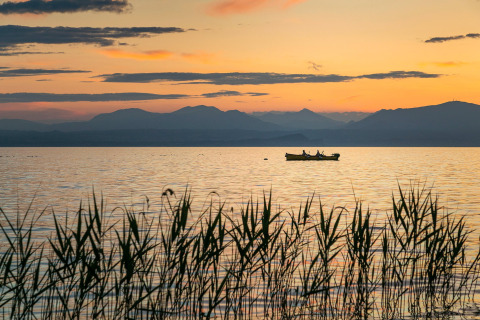Solnedgang ved Camping Cisano/San Vito, Italien, med en båd på søen og bjerge i baggrunden.