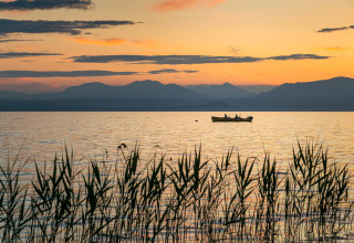 Solnedgang ved Camping Cisano/San Vito, Italien, med en båd på søen og bjerge i baggrunden.