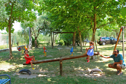 Niños juegan en un balancín en un parque infantil sombreado, vigilados por un adulto, árboles y coche cerca.