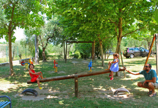 Children play on a seesaw in a shaded playground, watched by an adult, with trees and parked car nearby.