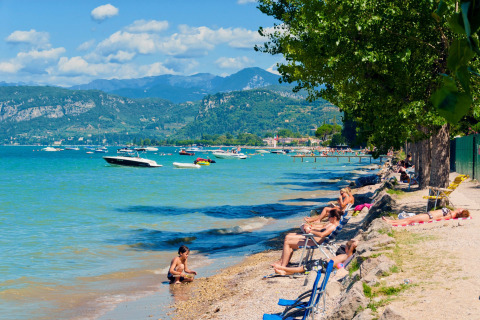 Villaggio turistico sul lago con bagnanti, barche sull’acqua e montagne sullo sfondo in Veneto, Italia.