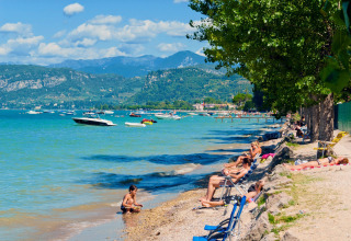 Holiday park by a lake with sunbathers, boats on the water, and mountains in the background in Veneto, Italy.