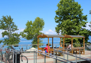Persone sedute sotto tettoie in legno al Camping Cisano/San Vito in Veneto, Italia, vista lago.